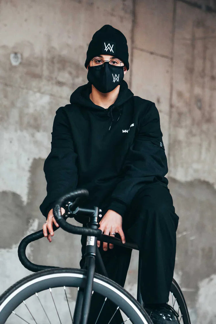 Urban-style portrait of a person seated on a bicycle wearing a coordinated Alan Walker outfit consisting of a black hoodie, face mask, and beanie, all featuring the distinctive 'AW' logo, set against a concrete wall for an edgy look.