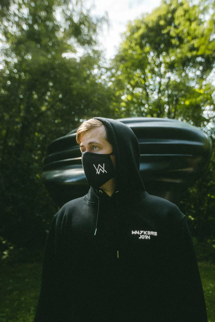 Outdoor portrait of a person clad in Alan Walker's signature merchandise, featuring the Core Logo Hoodie and Face Mask, both adorned with the 'AW' logo, set against a lush greenery backdrop.