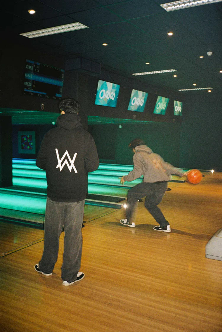 Two individuals at a bowling alley, one in the foreground wearing the Alan Walker Core Logo Hoodie with a prominent white 'AW' logo on the back, capturing a casual and fun social moment.