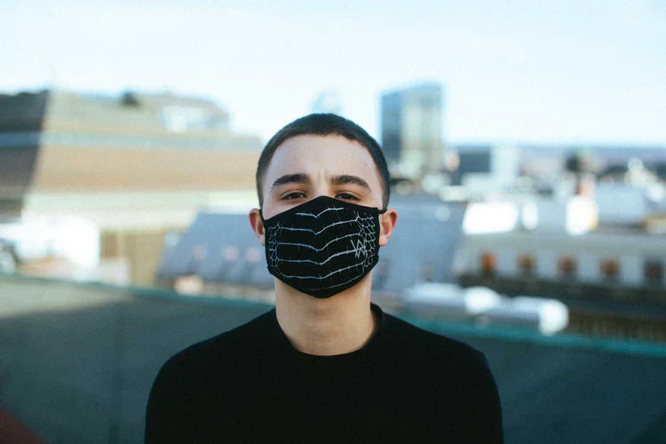 Young man standing on a rooftop, wearing a black face mask with a reflective spider web design, urban skyline in the background, highlighting a blend of safety and contemporary style.