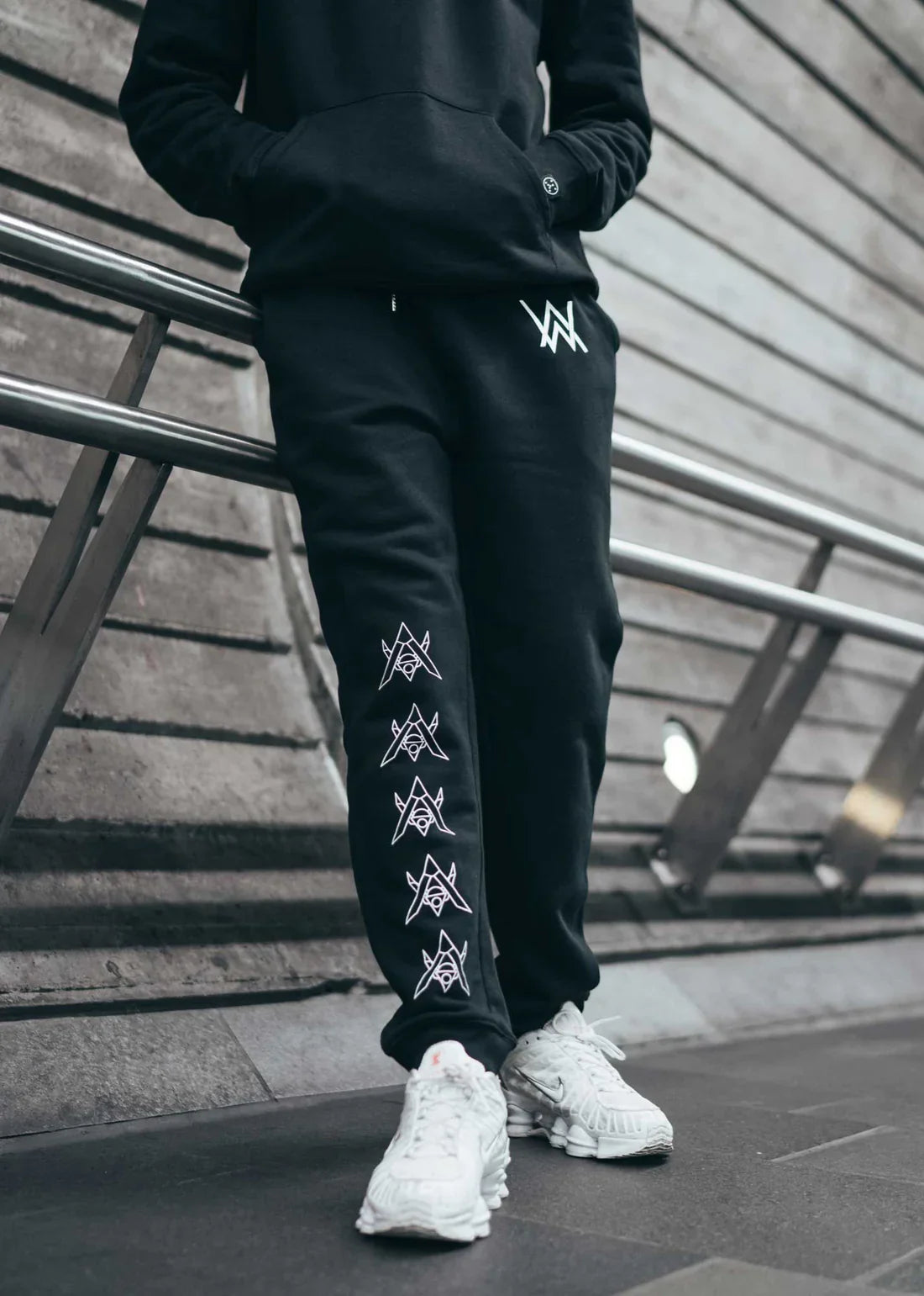 Person posed on stairs wearing Alan Walker Core Drone Sweatpants with distinctive white drone graphics down the right leg and a classic 'AW' logo on the left hip, paired with white sneakers for a casual urban look.