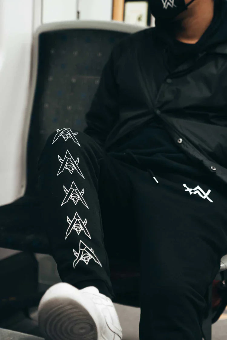 Person casually seated, sporting Alan Walker Core Drone Sweatpants with a unique white drone graphic design along the leg, paired with a black hoodie and white sneakers, exuding a relaxed urban vibe.