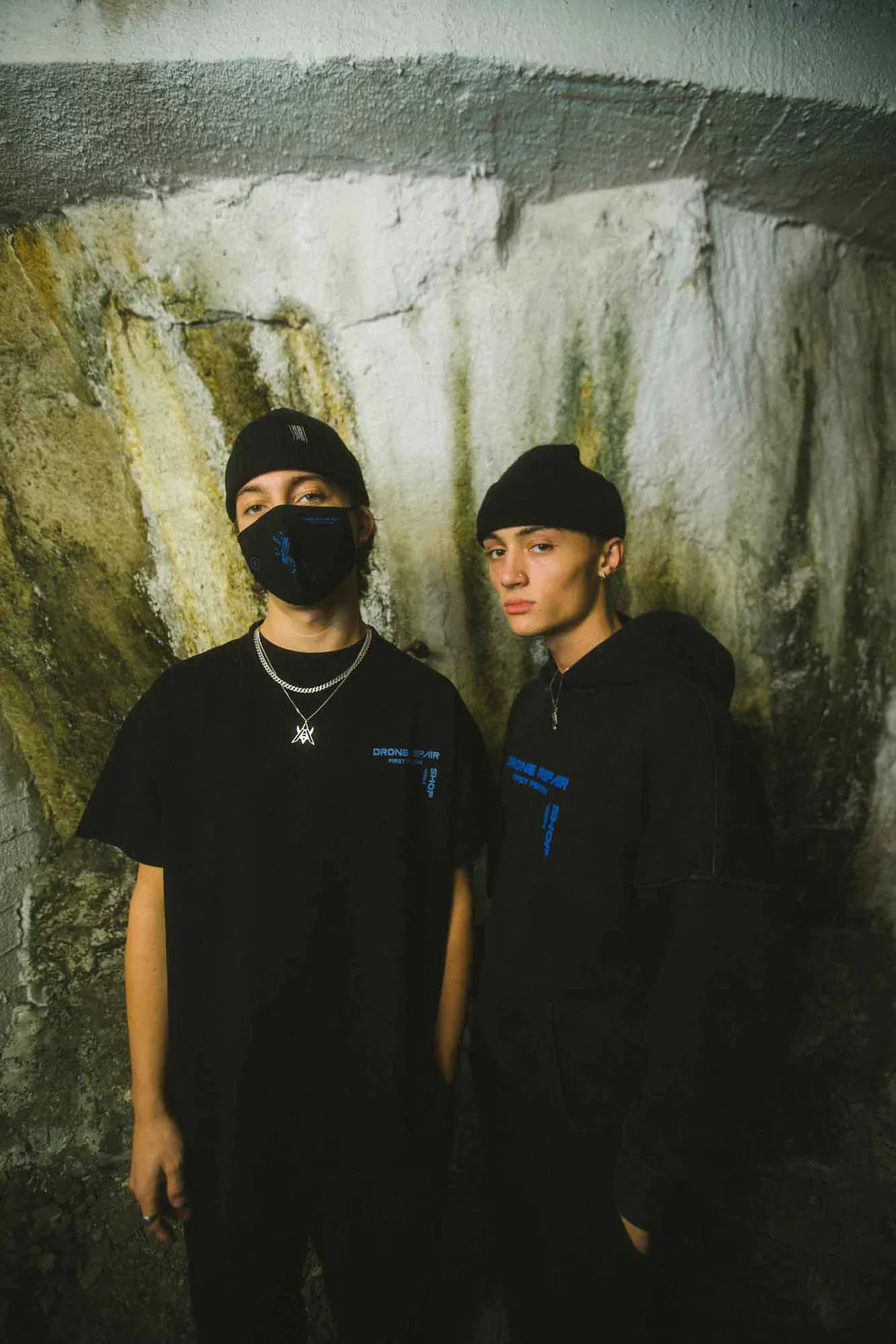 Two individuals standing against a mottled wall, one wearing a black Drone Repair Shop Mask with blue design, a black T-shirt, and the other in a black hoodie and beanie from the same collection, both with urban styling.