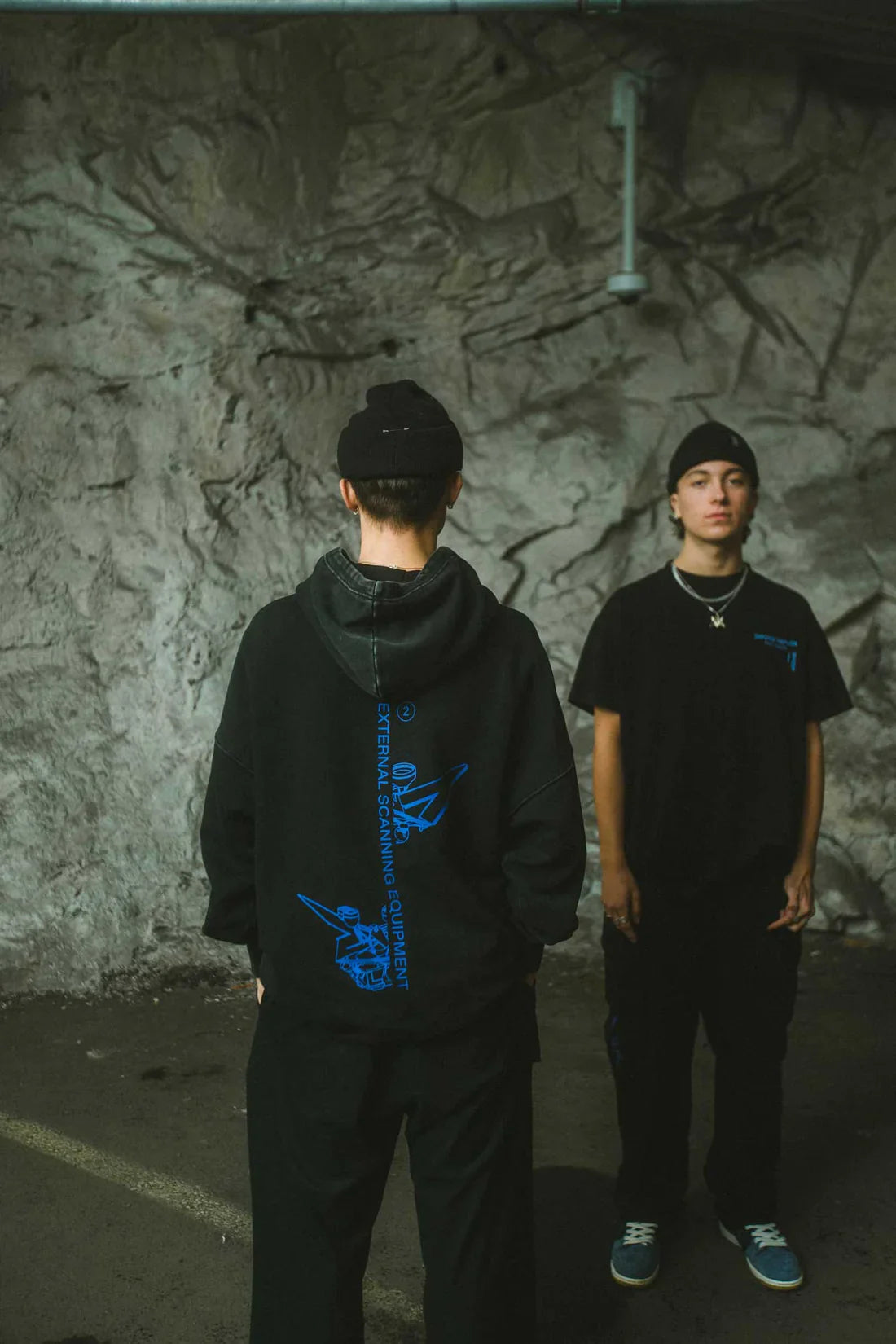 Two individuals in an urban setting, one facing away showcasing the back graphic of the black Drone Repair Shop Hoodie with blue design, and the other facing forward, both wearing beanies and casual attire.