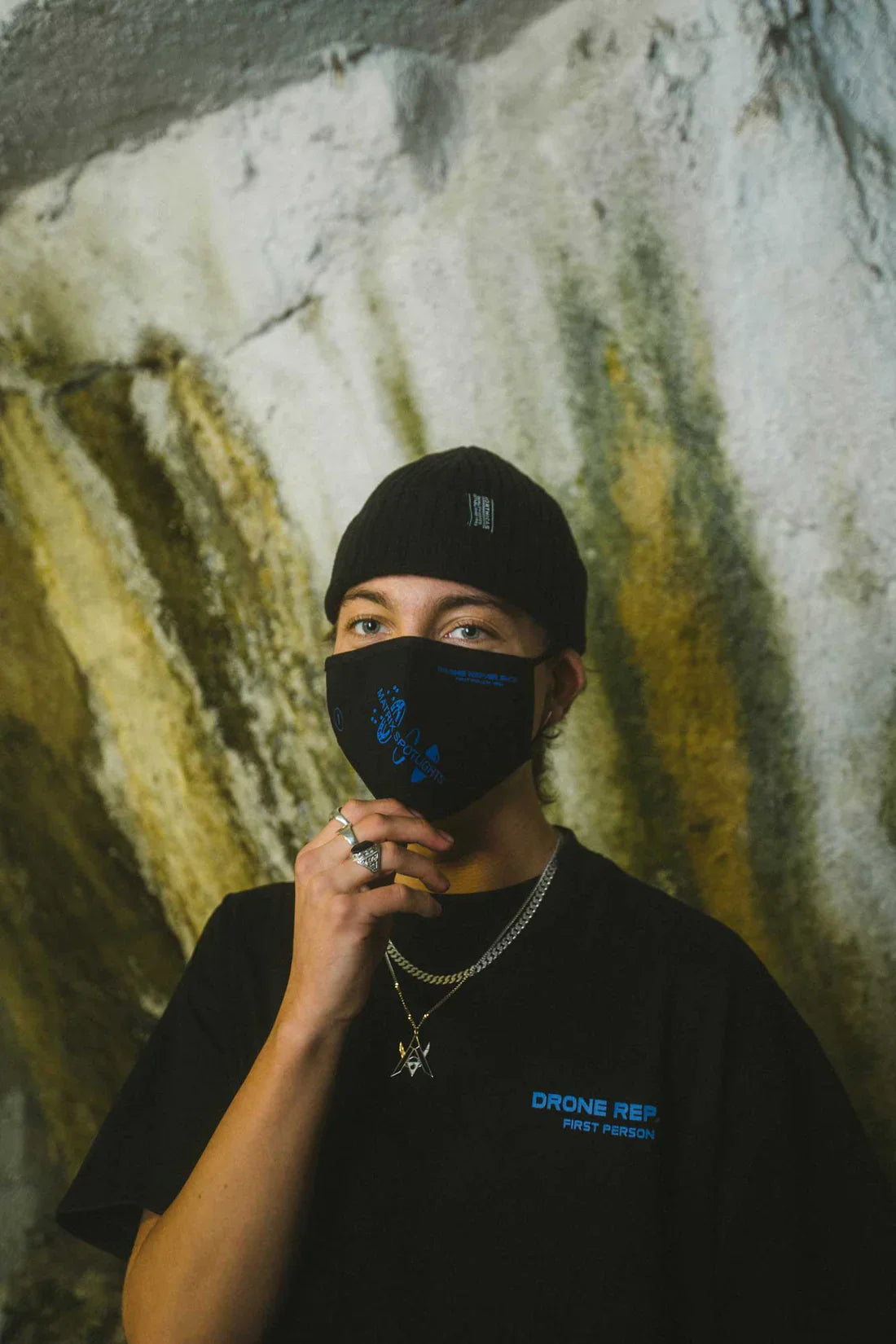 Individual with a black beanie and wearing the Drone Repair Shop Mask in black with blue graphics, paired with a matching black T-shirt, set against a textured wall backdrop.