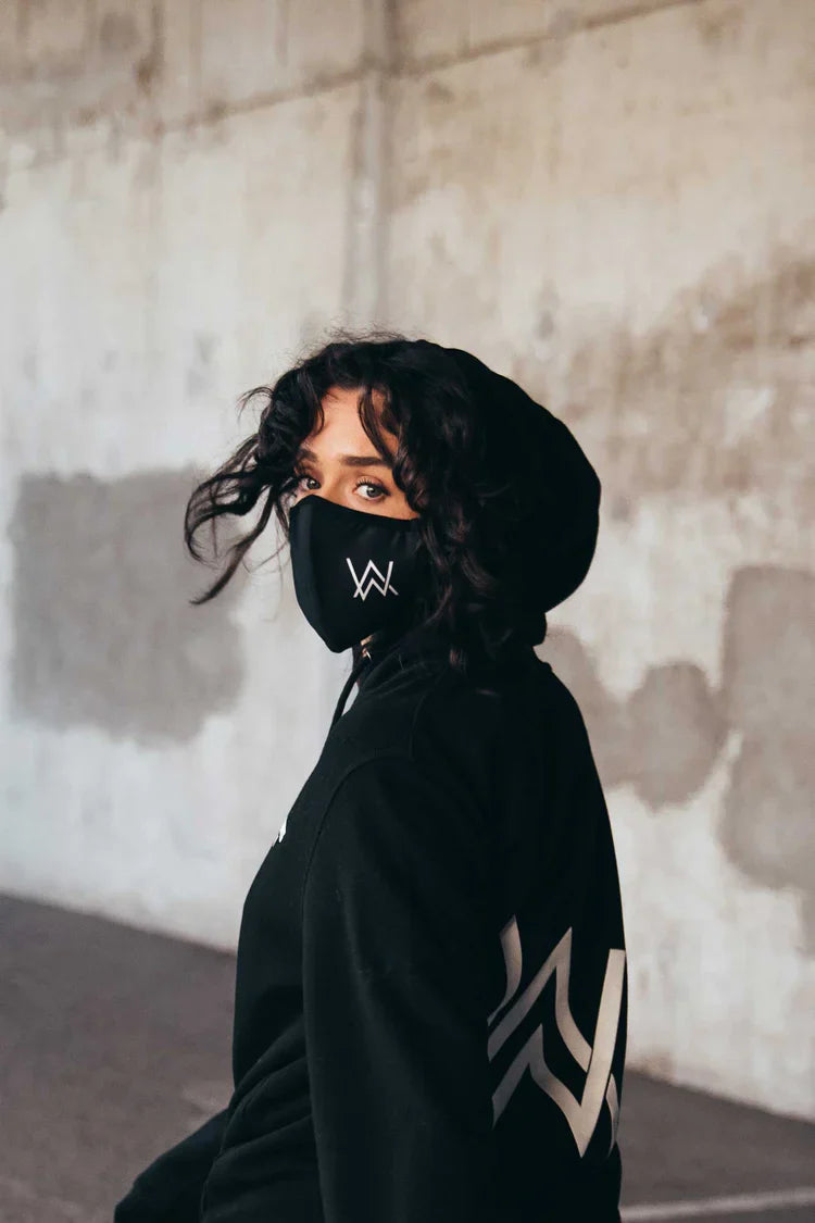 Side profile of a person wearing the Alan Walker Core Logo Face Mask and black hoodie with large 'AW' logo, giving a modern and moody look against an industrial concrete wall background.