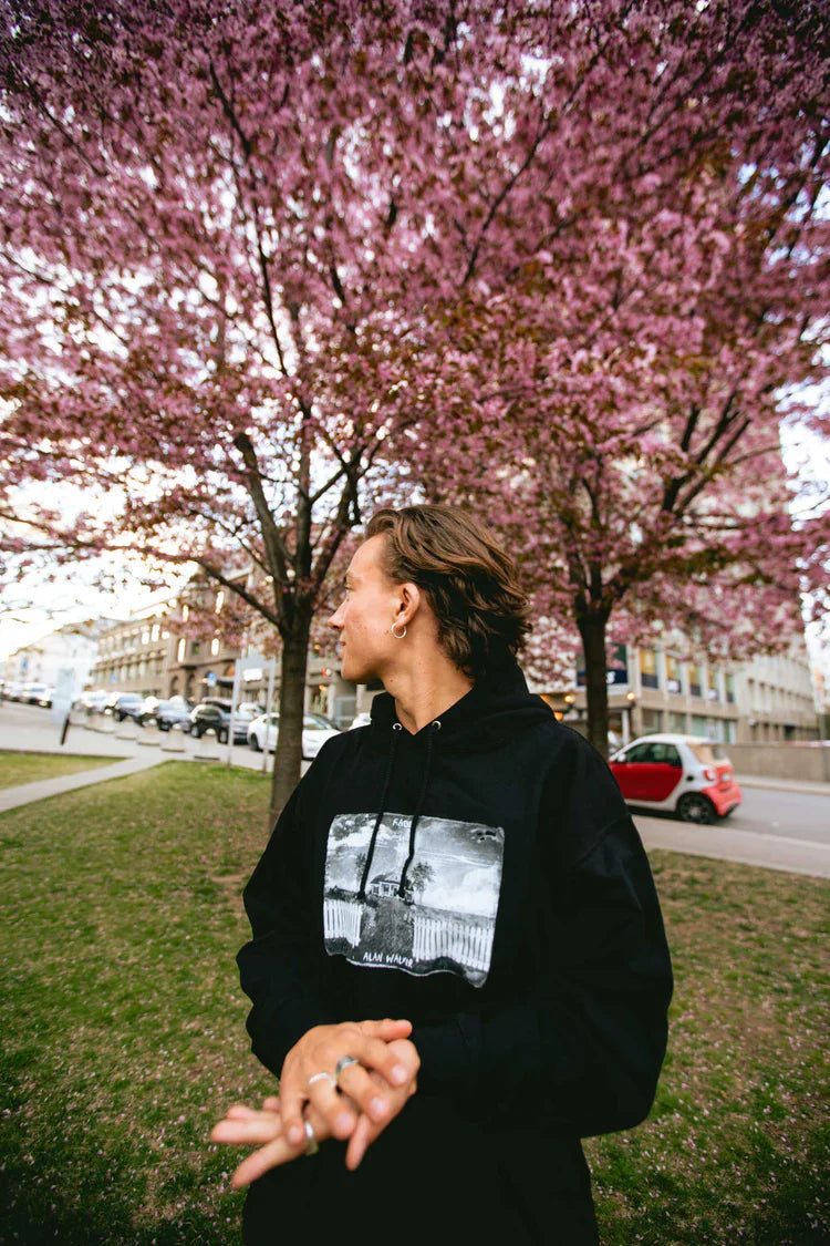 Person outdoors wearing Alan Walker's Faded 2.0 Hoodie with a central graphic, standing in profile against a backdrop of pink cherry blossoms.