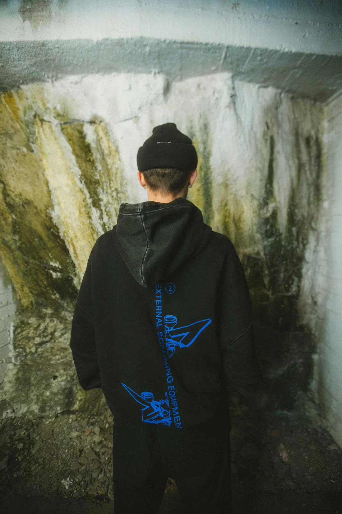 Rear view of a person wearing a black beanie and black Drone Repair Shop Hoodie with a distinctive blue graphic on the back, standing in front of a textured wall with natural discoloration.