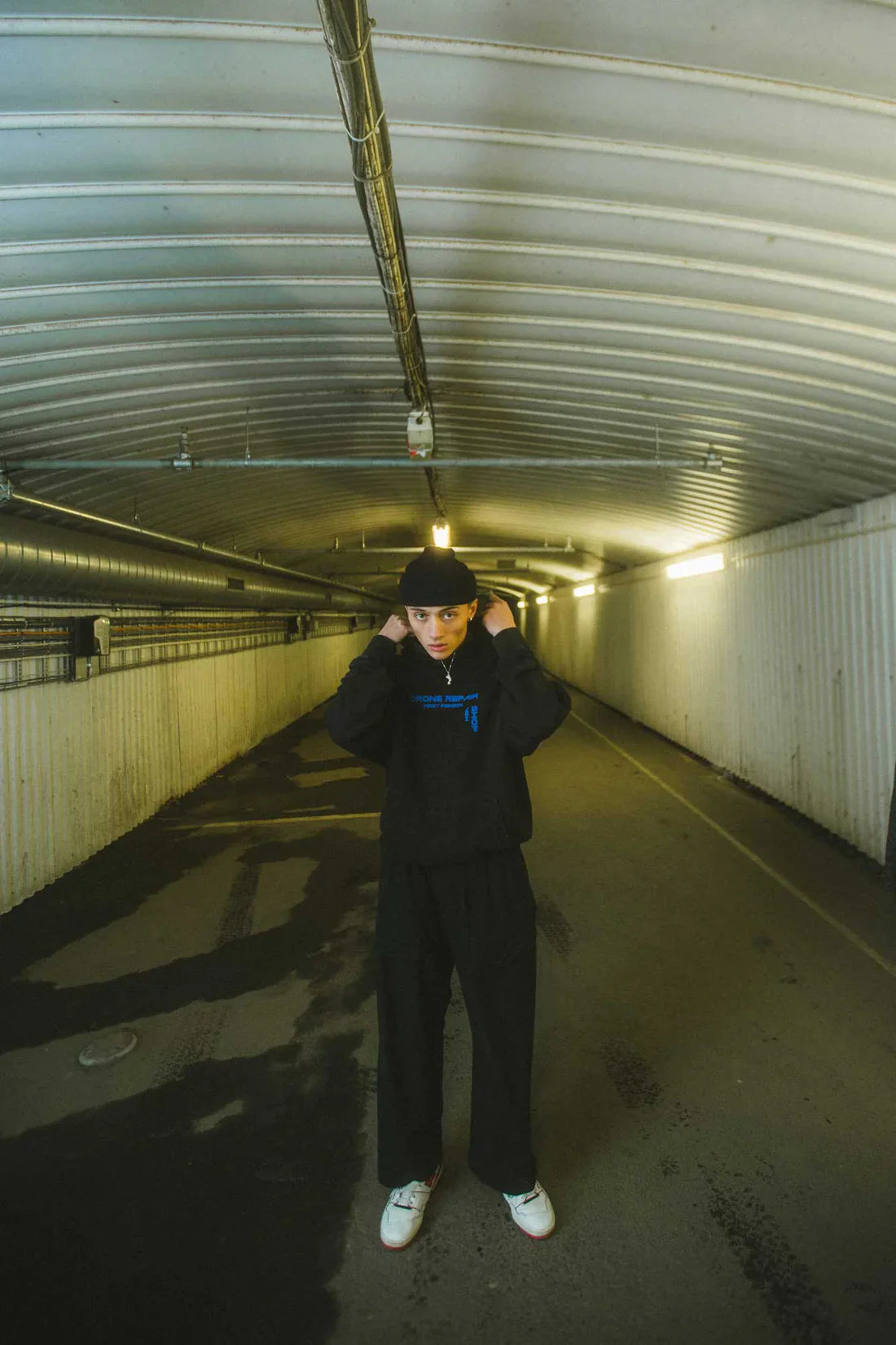 Person standing in a tunnel adjusting a black beanie, wearing the black Drone Repair Shop Hoodie with blue logo, complemented by casual black pants and white sneakers.