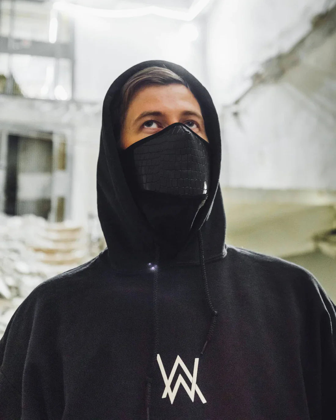 Close-up of a person wearing Alan Walker's signature black hoodie with the 'AW' logo, paired with a textured black face mask, set against an industrial interior background.