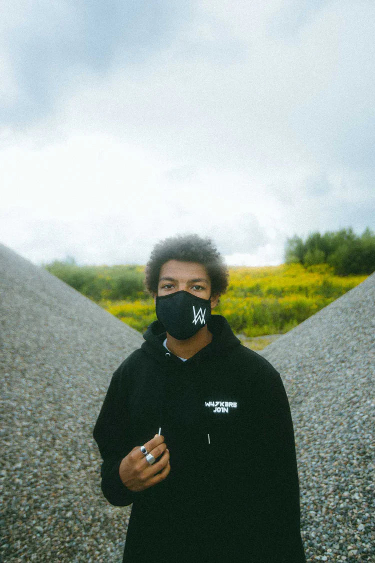 Individual in a natural setting wearing Alan Walker merchandise, including a black hoodie with 'W4LK3RS JOIN' text and a face mask featuring the 'AW' logo, embodying a spirit of unity and music culture.