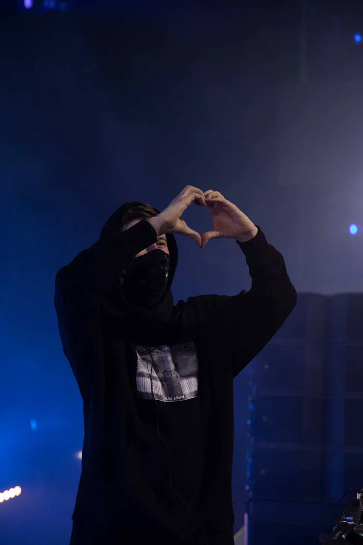 Artist on stage forming a heart shape with hands, wearing Alan Walker's Faded 2.0 black hoodie featuring a central cityscape graphic, in a concert setting with moody blue stage lighting.