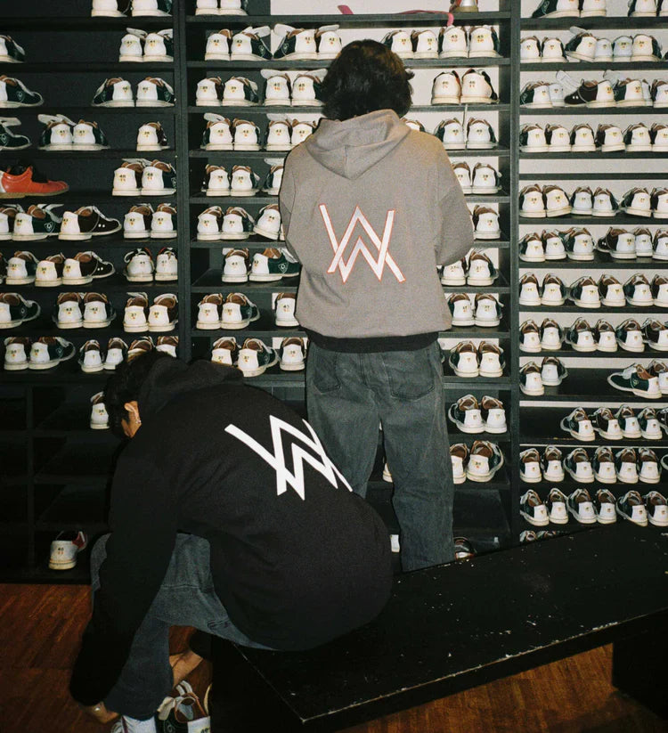 Two individuals at a shoe rental counter in a bowling alley, one standing and one crouching, both wearing Alan Walker Core Logo Hoodies with the distinctive 'AW' logo, surrounded by rows of bowling shoes on shelves.