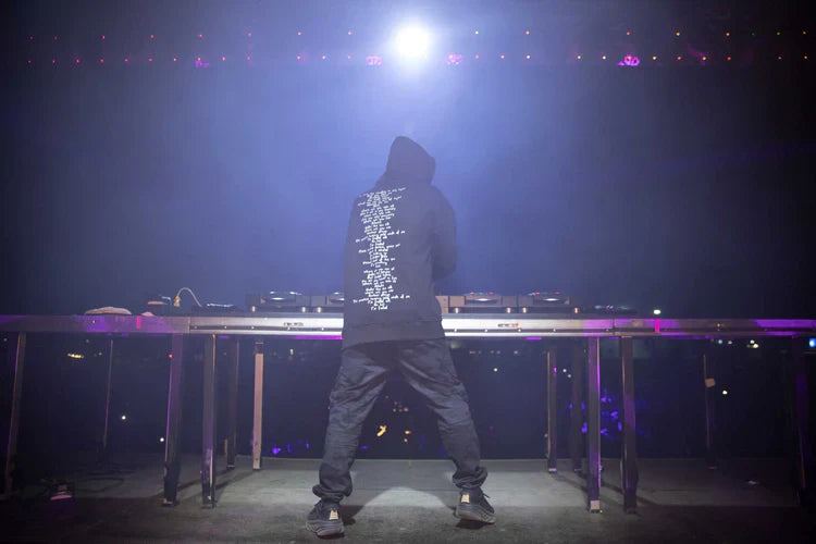 Rear view of a DJ on stage wearing Alan Walker's Faded 2.0 black hoodie with song lyrics printed on the back, facing a mixing console under vibrant purple stage lights.