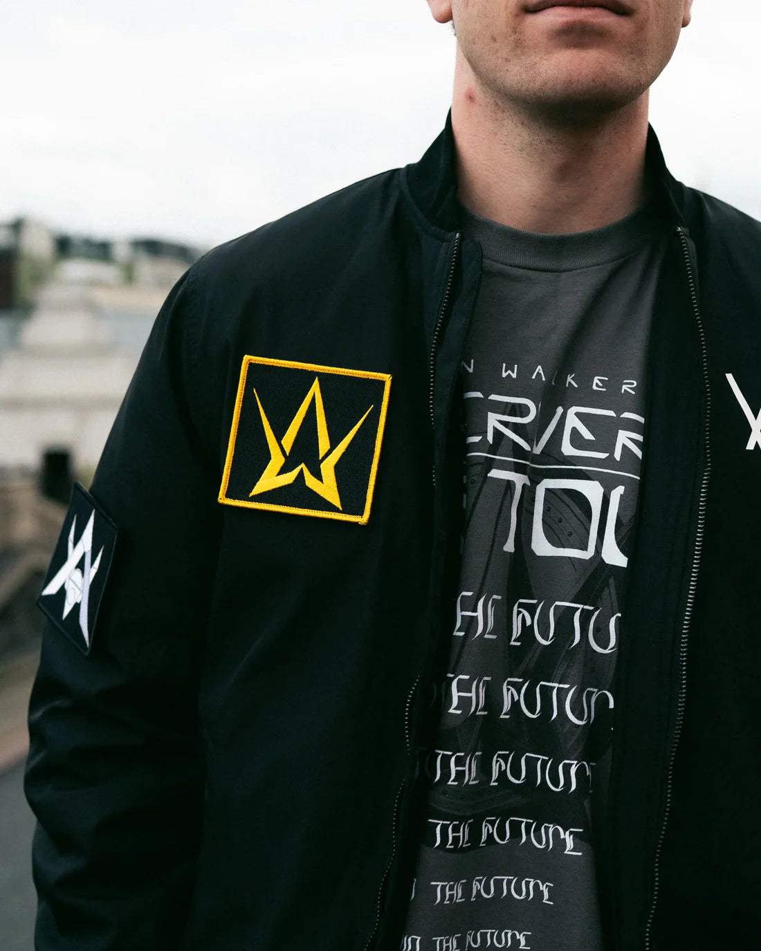 Man wearing a black bomber jacket with a large golden Alan Walker logo on the left sleeve and a smaller white logo on the right, over a T-shirt with Alan Walker's repeated 'Different World' album text.
