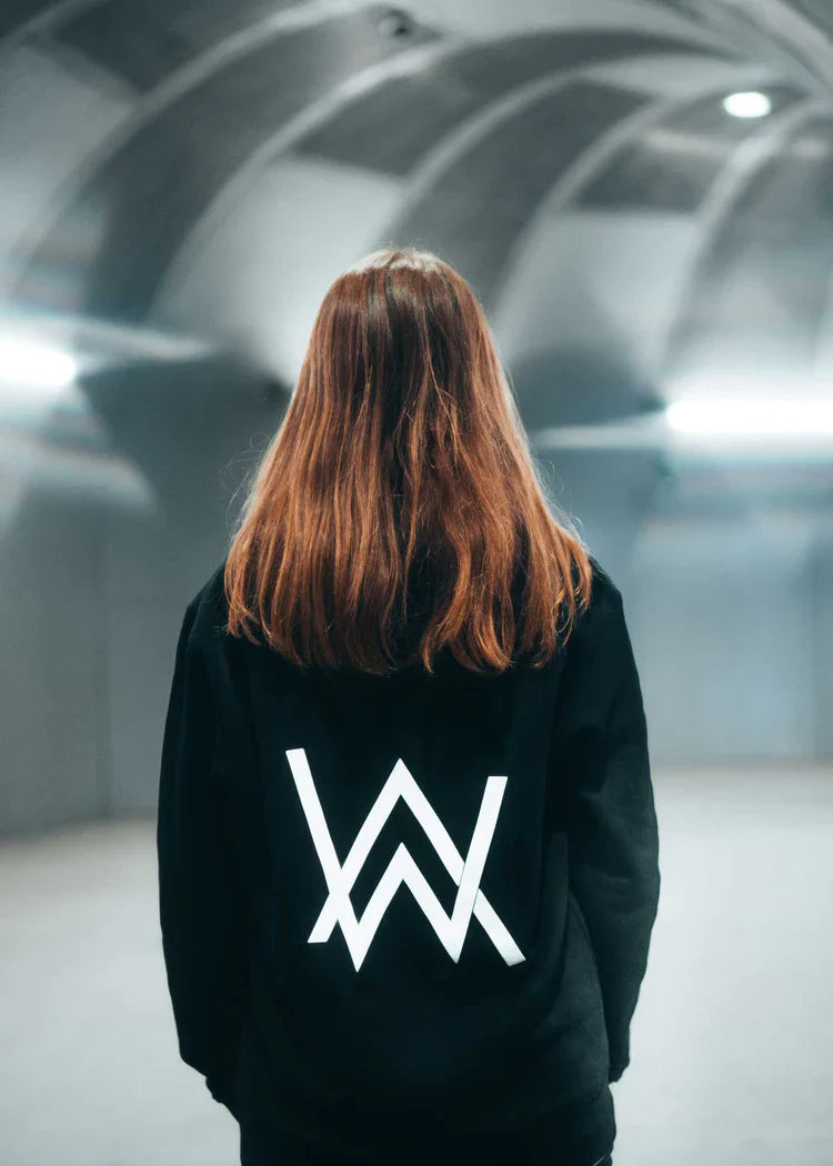 Back view of a person with long hair wearing the Alan Walker Core Logo Hoodie, featuring a large white 'AW' emblem, in a modern architectural environment with a futuristic aesthetic.