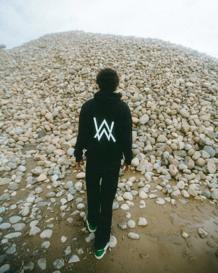 Individual ascending a pebble hill wearing the CORE LOGO ZIP HOODIE with a striking 'AW' emblem on the back, harmonizing with the raw, earthy environment, under a muted sky.