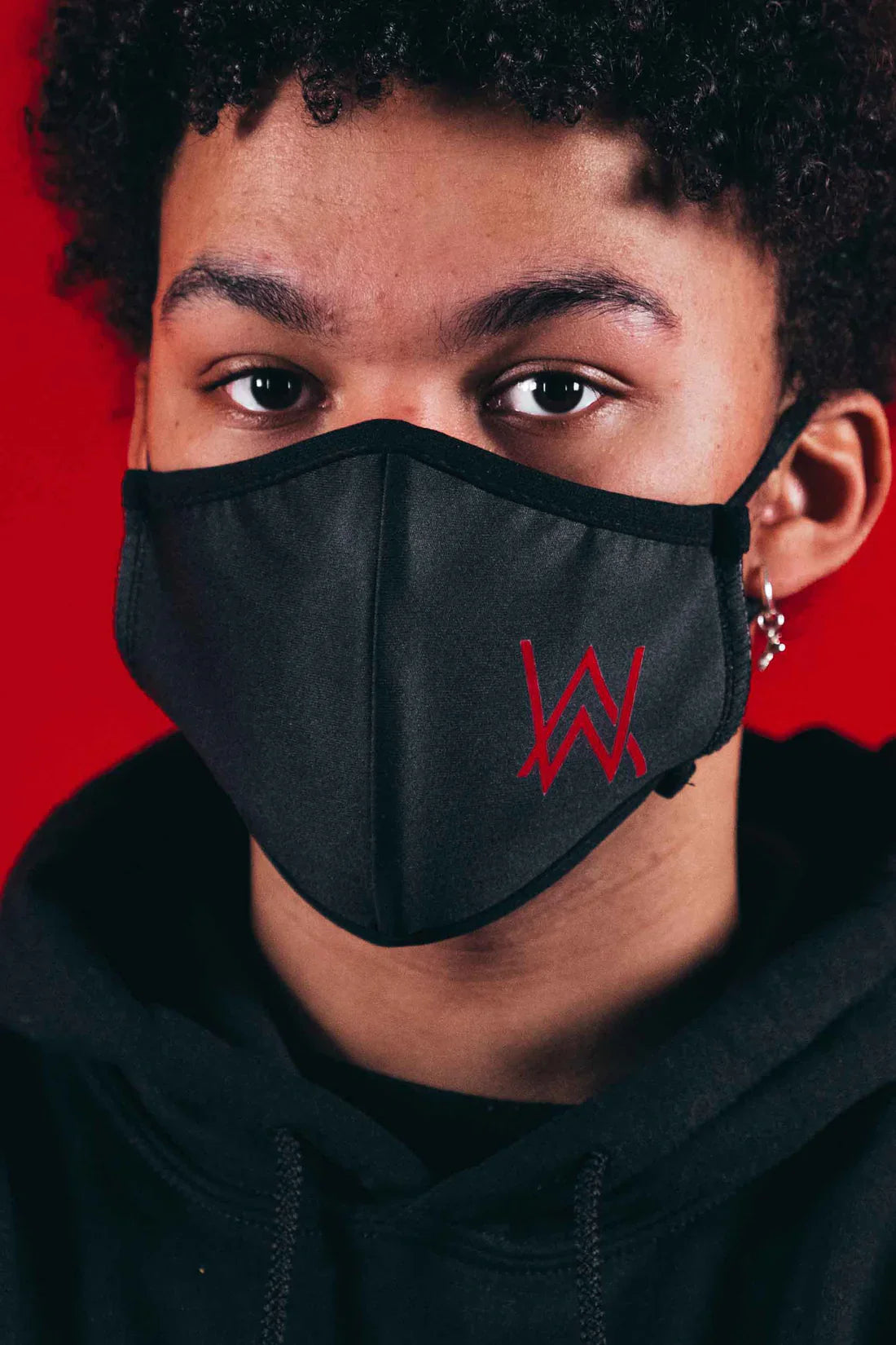 Young person wearing a black protective mask with a bold red Alan Walker logo, set against a red backdrop.