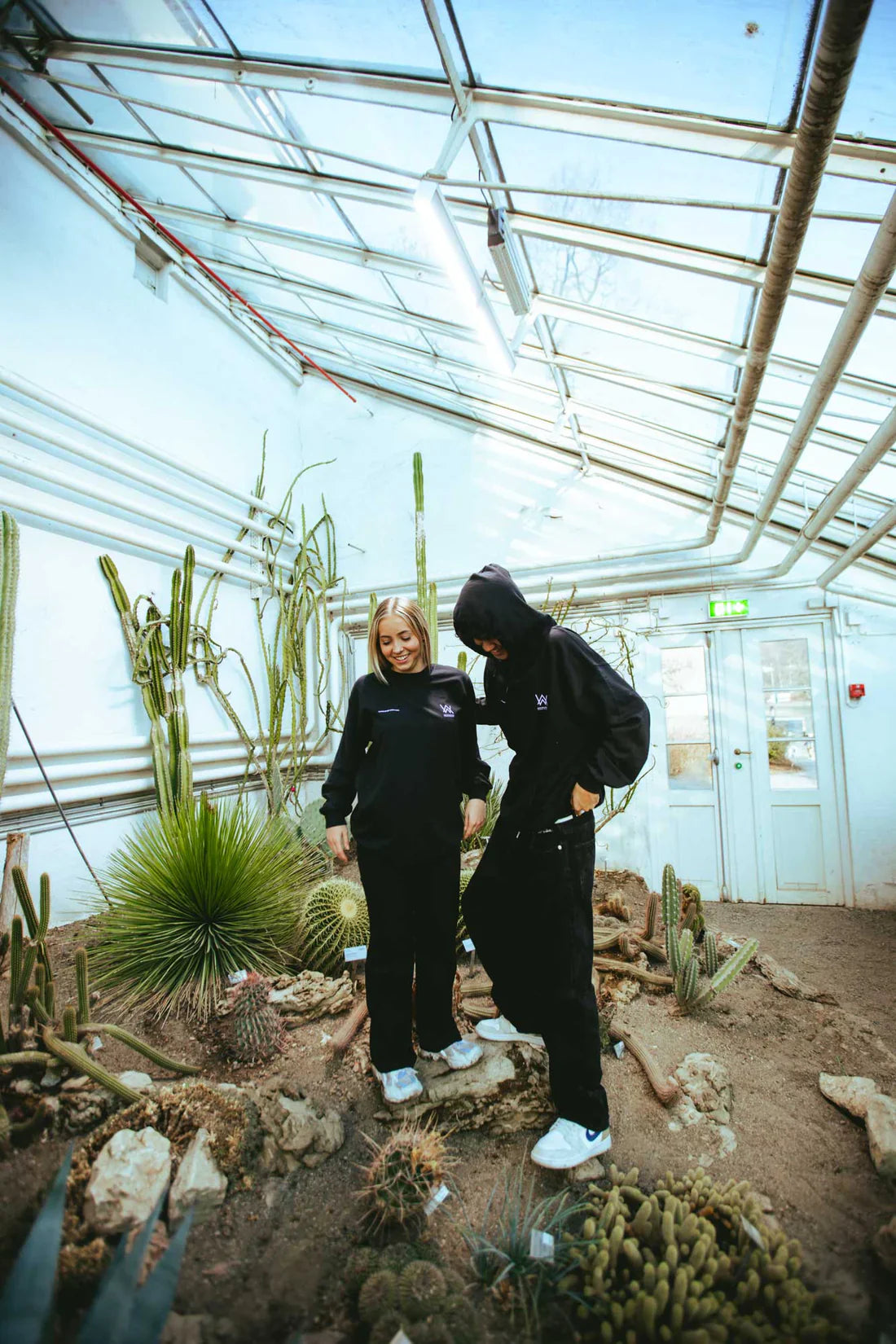 A duo in Melting Rose Hoodies showcasing the unity of Alan Walker fans, set against the backdrop of a serene greenhouse.