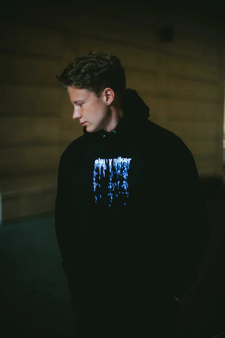 Profile view of a man in a dimly lit setting, wearing an 'alan walker' digital rain graphic hoodie, resonating with the subdued tones of electronic music.