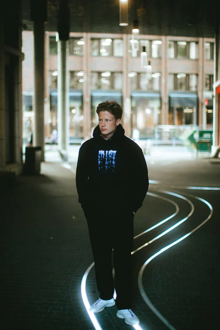 Nighttime urban shot of a man in a black hoodie with glowing blue 'alan walker' digital graphic, encapsulating city nightlife and electronic beats.