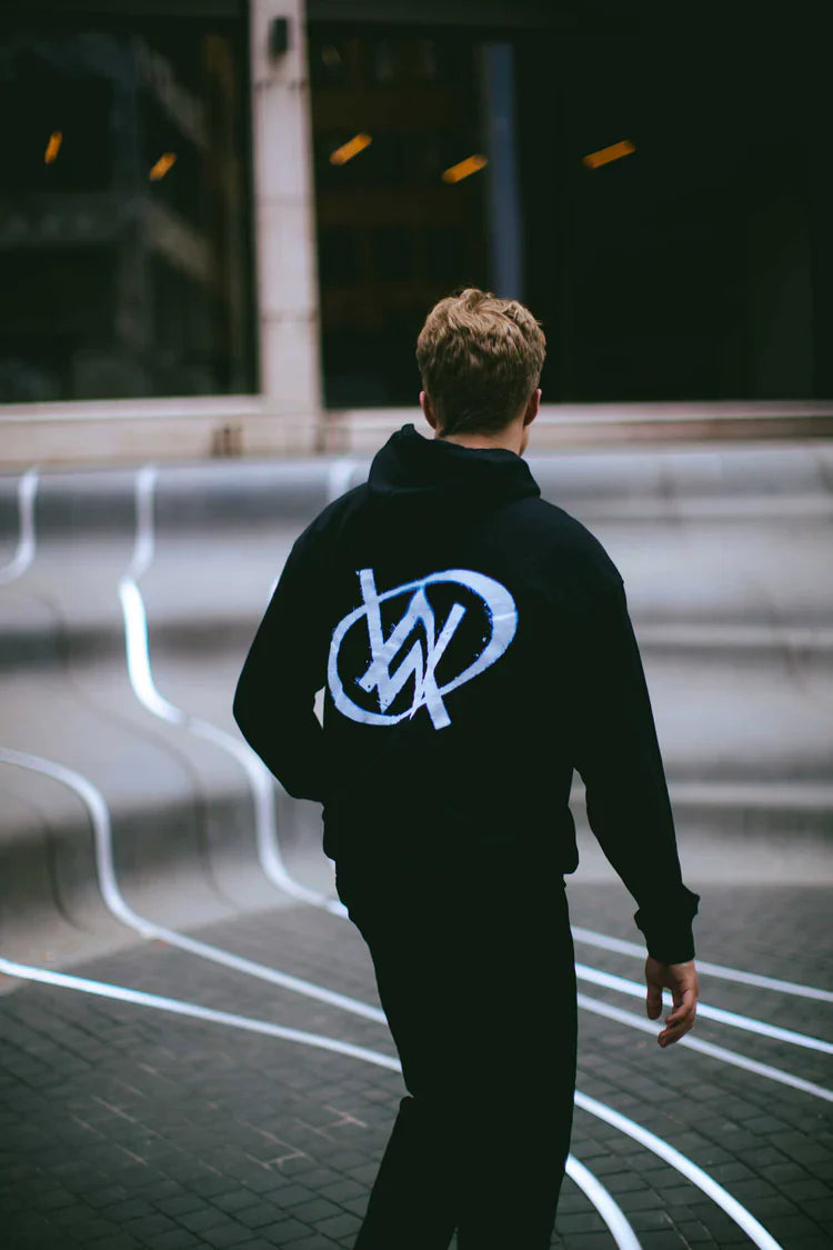 Man walking away on a city street, showcasing the iconic blue Alan Walker logo on the back of a black hoodie, embodying the rhythm of city life