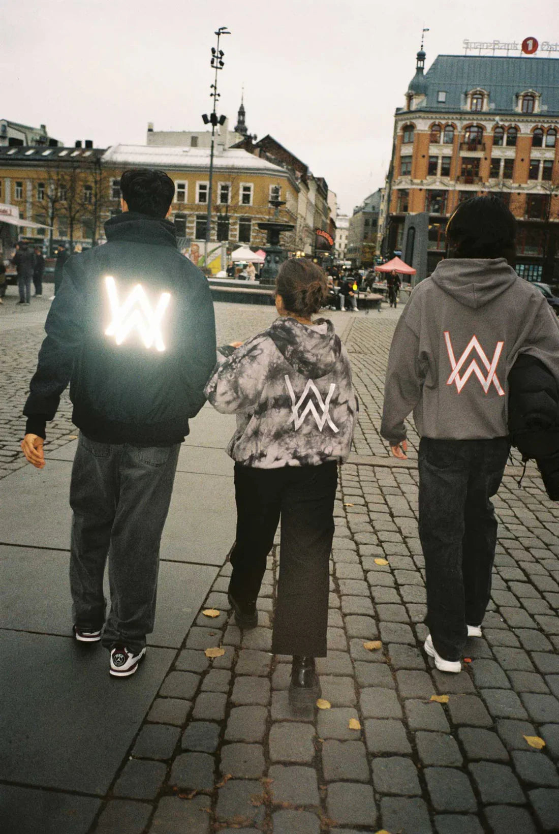 Three friends walking through a city square, one with a glowing Alan Walker logo on their jacket, flanked by two others in Alan Walker hoodies.