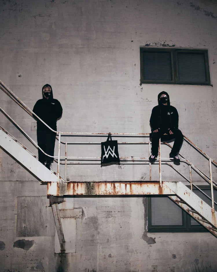 Two figures in Alan Walker's CORE LOGO ZIP HOODIES exude a rebellious urban vibe on a weathered staircase, with a CORE REFLECTIVE TOTE BAG draped over the railing, set against a stark industrial backdrop.