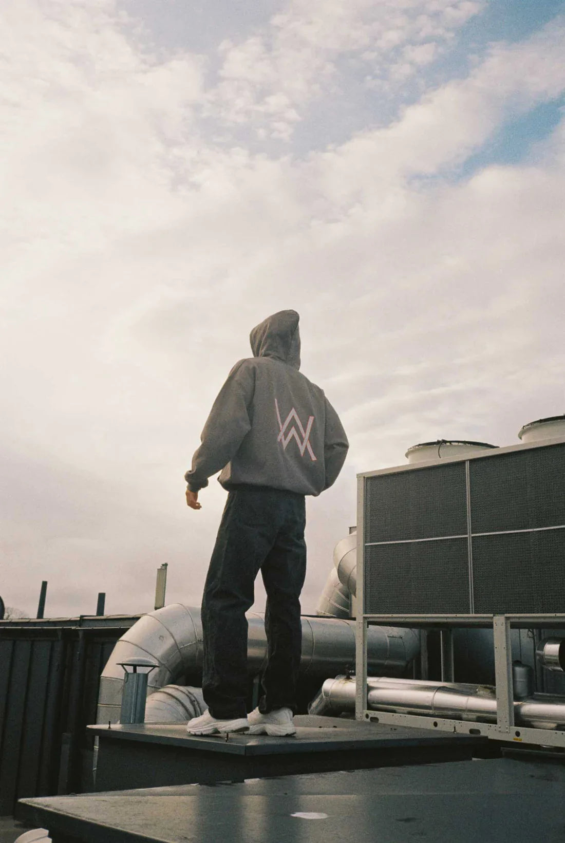 Person standing on a rooftop with their back turned, wearing Alan Walker's Gaming Hoodie featuring the iconic white Alan Walker logo against a cloudy sky backdrop.